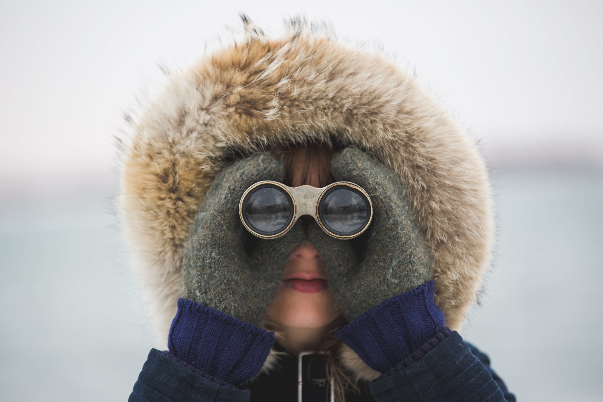 Child wearing a fur-trimmed hood and knitted gloves, looking through vintage binoculars.